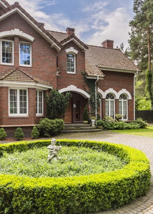 Round hedge and a small statue in the middle of a cobblestone driveway to an elegant, english style house with white windows and brick exterior